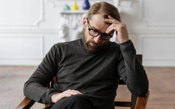 Um homem com roupas elegantes, óculos de grau e barba, e pensativo durante a sessão de terapia com o psicólogo Davi Scharanck.